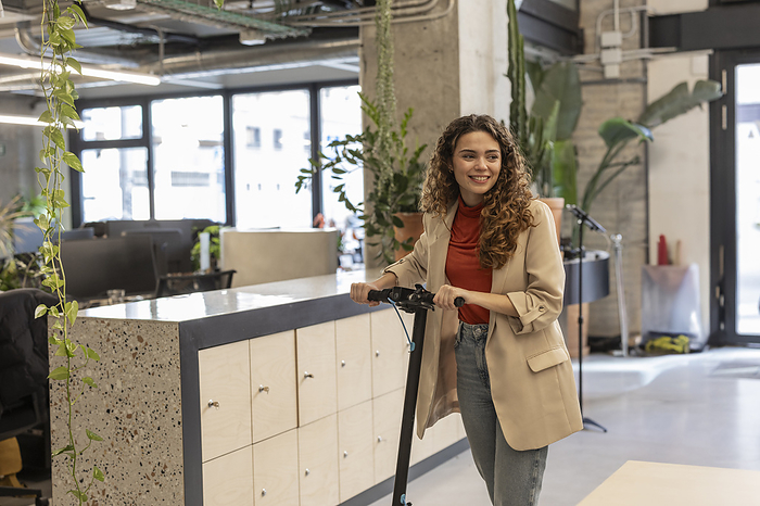 Smiling young businesswoman riding electric push scooter at workplace