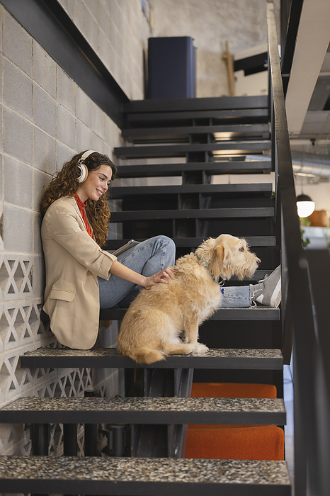 Smiling businesswoman listening to music sitting with dog on staircase at office