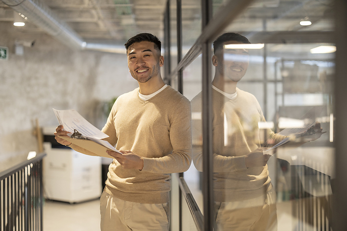Smiling young businessman with documents leaning on glass wall at office