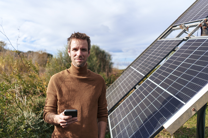 Smiling man standing with smart phone near solar panel on sunny day