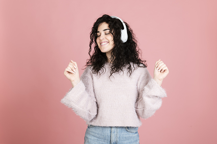 Happy young woman wearing wireless headphones listening to music against pink background