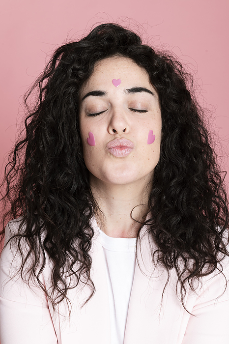 Woman puckering with heart shaped stickers on face against pink background