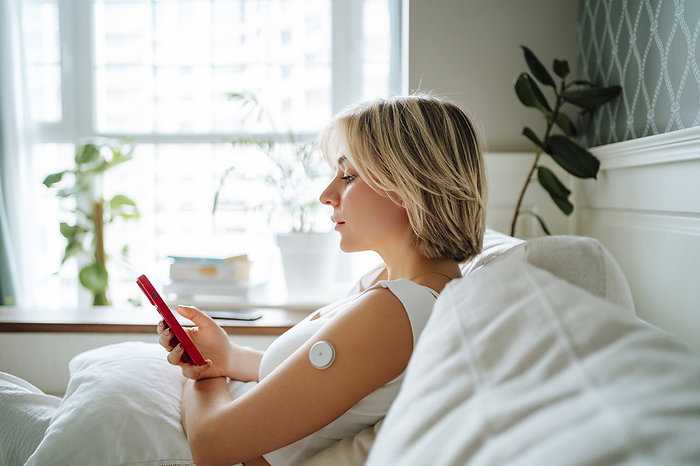 Woman using smart phone with glucose sensor on arm at home