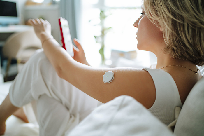 Glucose measurement sensor attached on woman's arm using smart phone on sofa at home