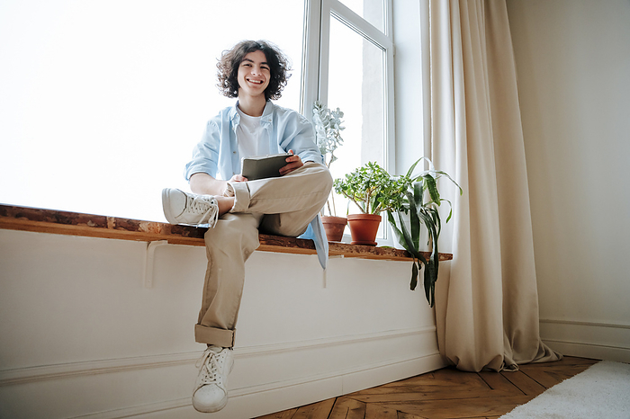 Smiling young man sitting with tablet PC on window sill at home