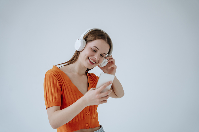 Smiling woman wearing wireless headphones and using smart phone in front of white background