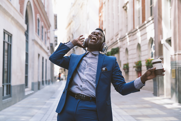 Cheerful mature businessman wearing wireless headphones holding coffee cup and enjoying on street