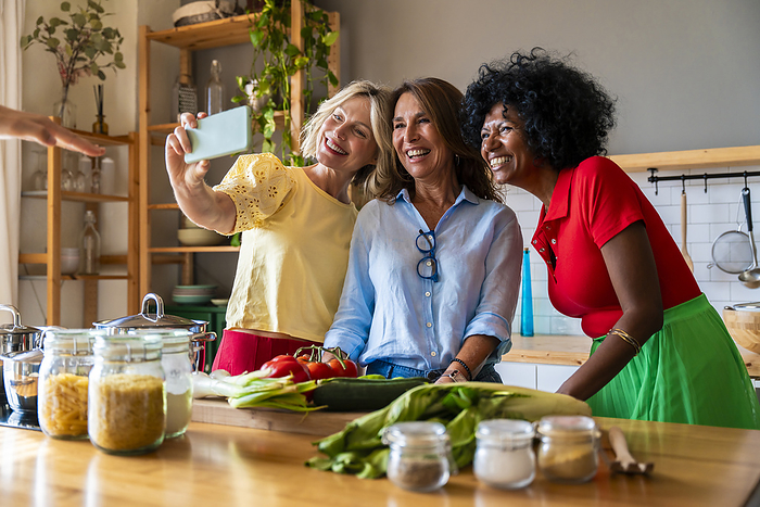 Happy multiracial women taking selfie through mobile phone in kitchen at home