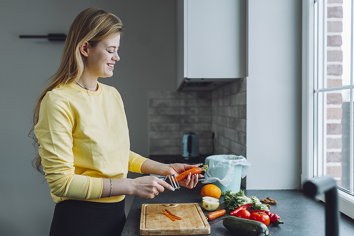 Smiling young woman peeling vegetables on kitchen counter at home