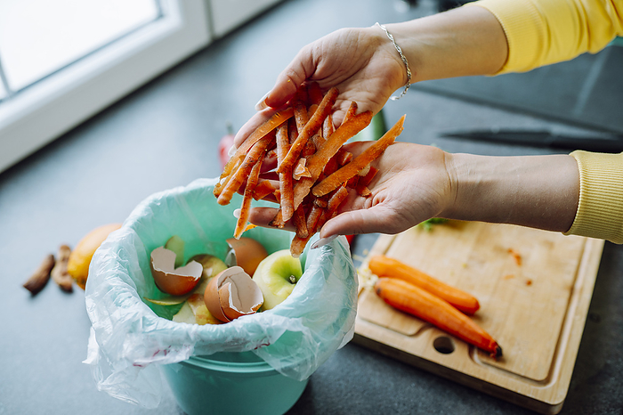Hands of young woman putting carrot peels in garbage can at home