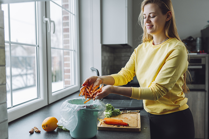 Smiling woman putting carrot leftovers in garbage can at home