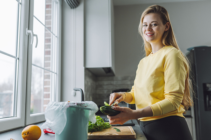 Smiling young woman peeling zucchini near kitchen counter at home