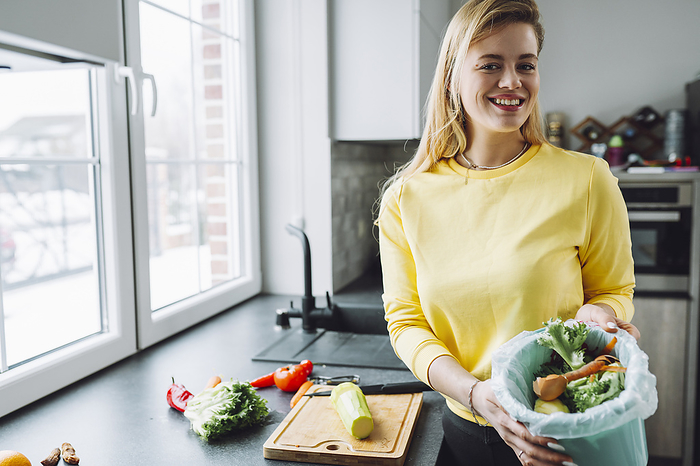 Smiling woman holding vegetables peels in garbage can at home