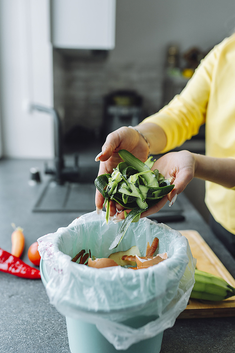 Young woman putting zucchini peels in garbage can at home