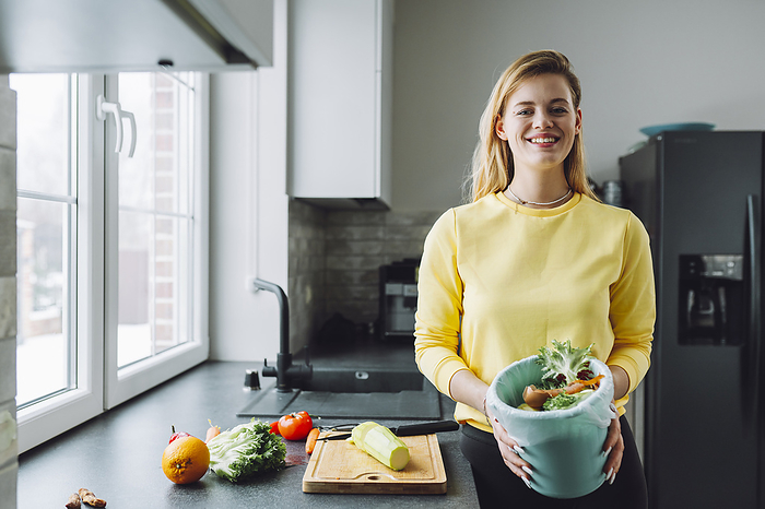 Smiling woman holding garbage can with vegetable peels at home