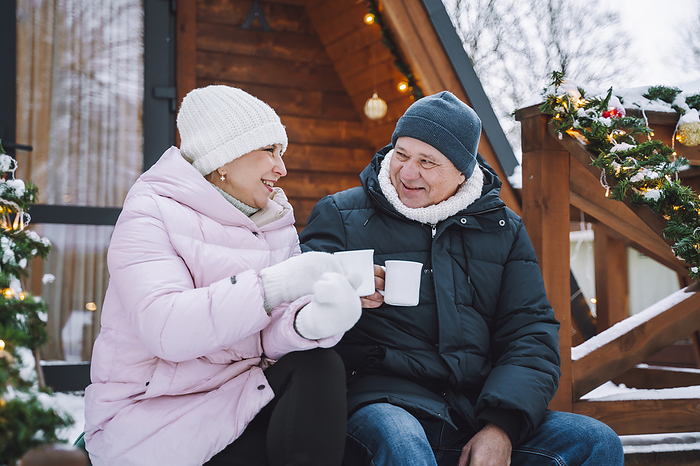 Happy mature couple enjoying hot tea sitting on porch near log cabin