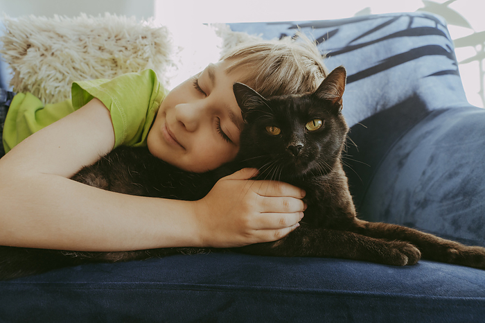 Smiling boy with eyes closed lying down with cat on sofa at home