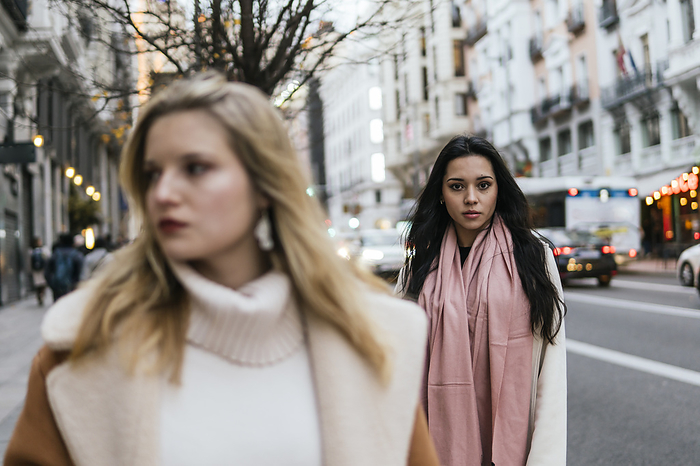 Serious woman wearing scarf and standing with friend on sidewalk in city