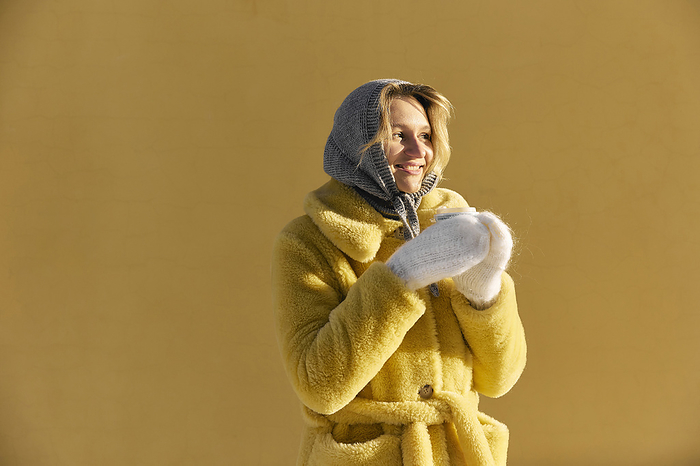 Happy young woman wearing fur coat and holding coffee cup in front of yellow wall