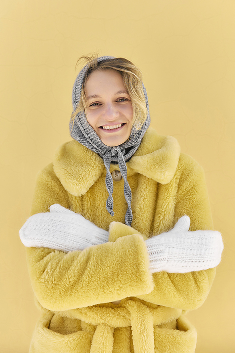 Happy young woman wearing fur coat and mittens feeling cold in front of yellow wall