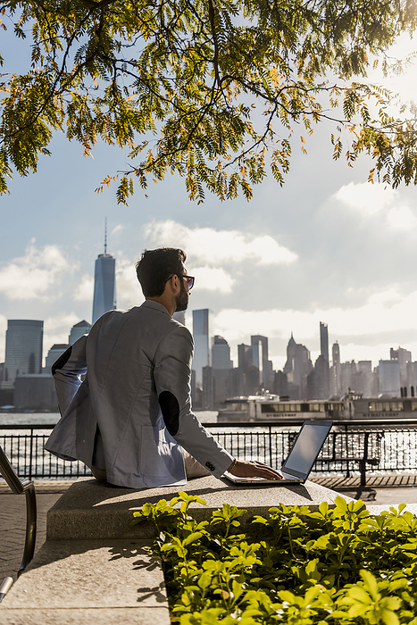 Businessman sitting on bench with laptop in city at sunny day