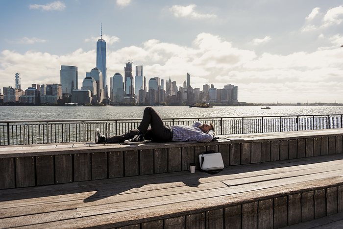 Carefree businessman taking nap lying on bench with skyline in background in city
