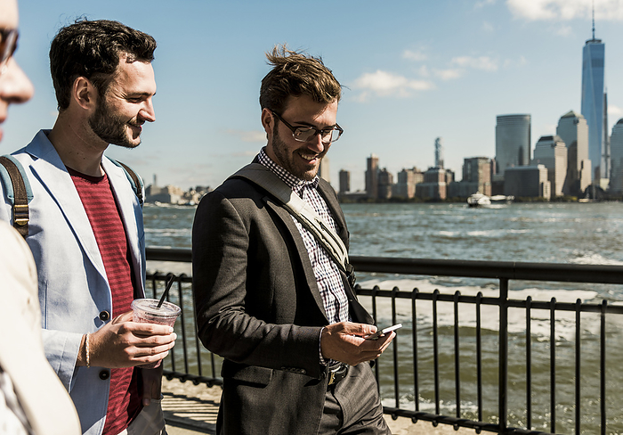 Happy businessmen with smart phone walking on promenade in city