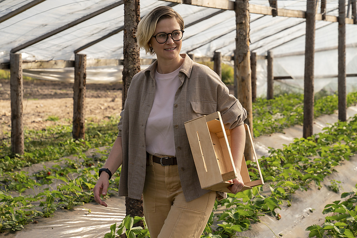 Smiling farmer holding crate and walking in greenhouse