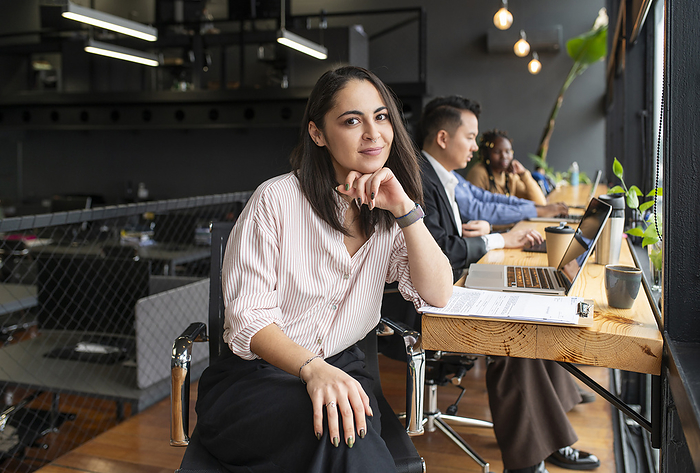 Smiling businesswoman with hand on chin sitting near colleagues at desk