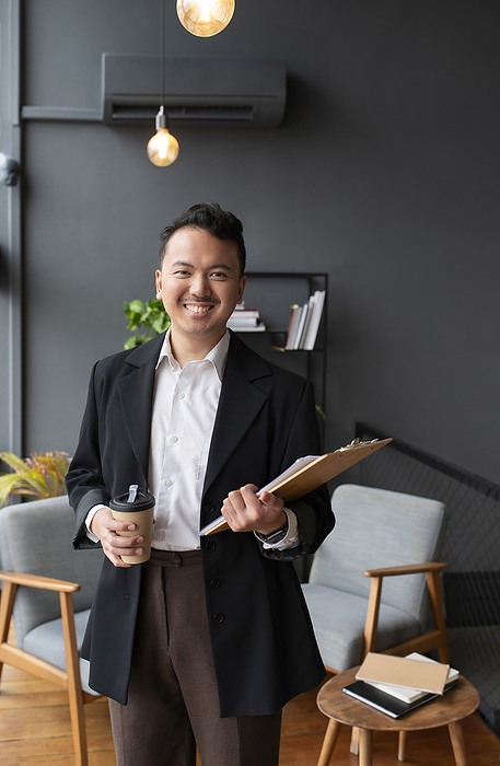 Smiling businessman standing with clipboard and coffee cup in office