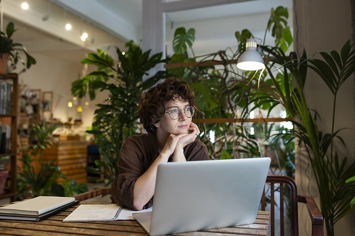 Thoughtful plant shop owner with laptop at table in store