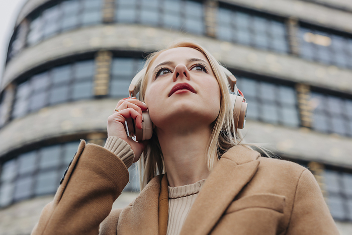 Contemplative woman listening to music through wireless headphones in front of building