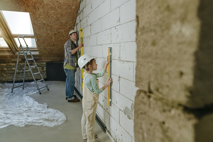Girl wearing hardhat and measuring wall through leveling tool with father at home