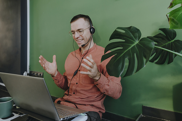 Smiling freelancer using headset and laptop for video call in front of green wall