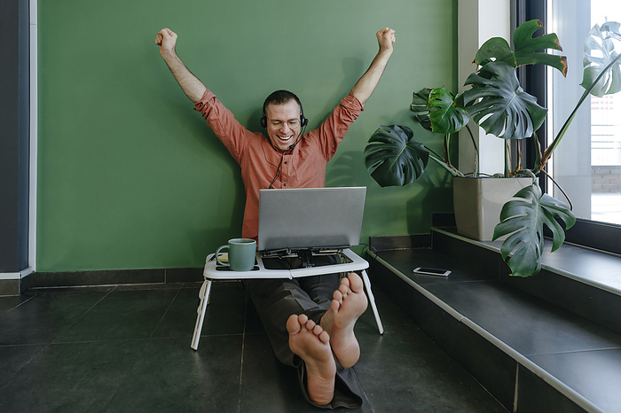Happy freelancer with arms raised cheering on video call in front of green wall