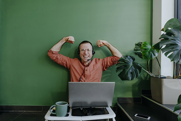 Happy freelancer flexing muscles near laptop in front of green wall