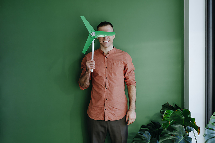 Smiling freelancer covering eyes with wind turbine model in front of green wall
