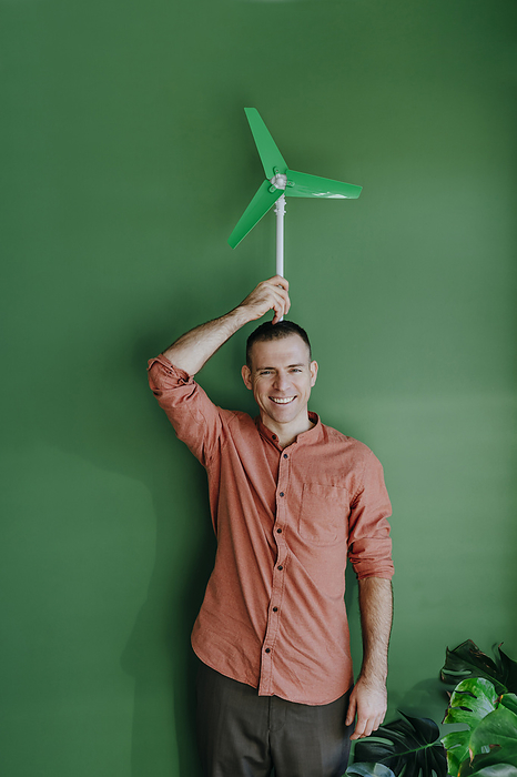 Happy freelancer holding wind turbine model on head in front of green wall