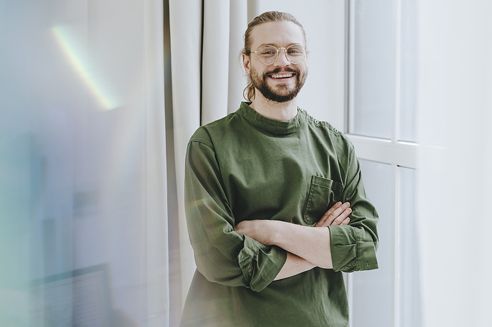 Happy freelancer standing with arms crossed near window at office