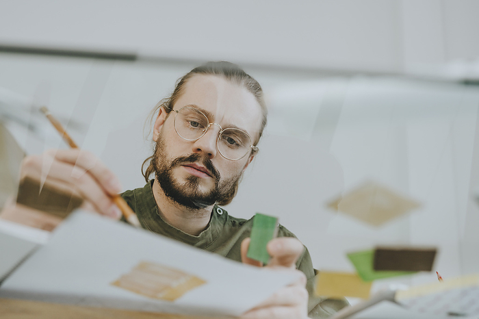 Focused furniture designer working at desk seen through glass at office