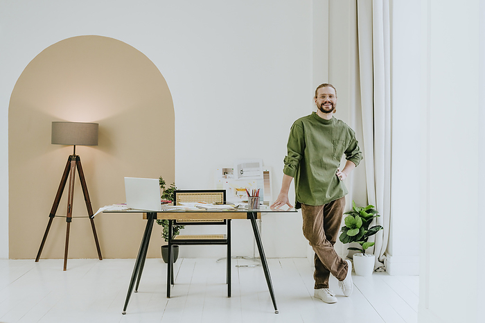 Happy interior designer standing with hand on hip near desk at office