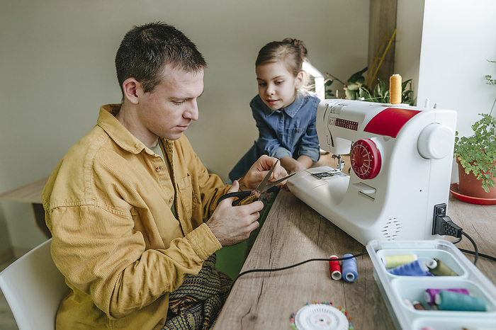 Daughter looking at father cutting needle with scissors by sewing machine at home