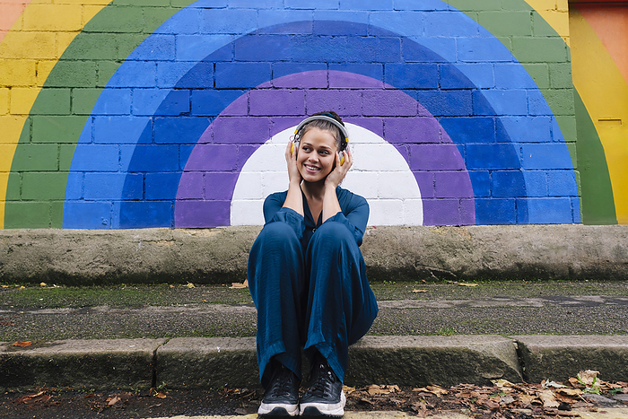 Smiling woman wearing wireless headphones and sitting on footpath in front of rainbow wall