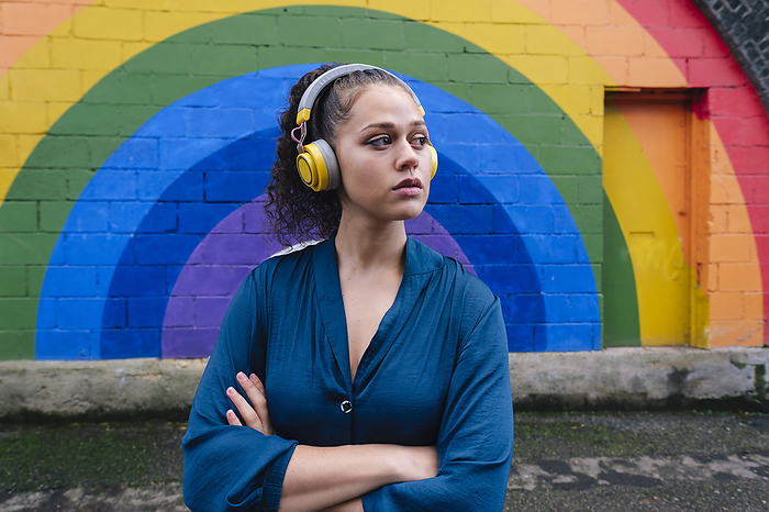 Young woman wearing wireless headphones and standing with arms crossed in front of rainbow wall