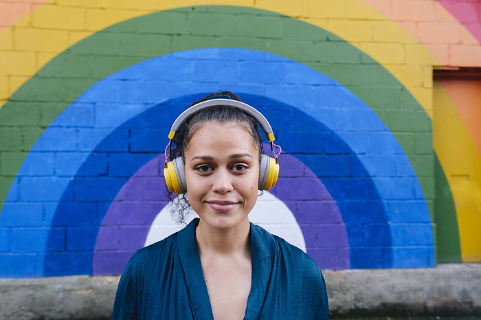 Smiling young woman wearing wireless headphones in front of rainbow wall