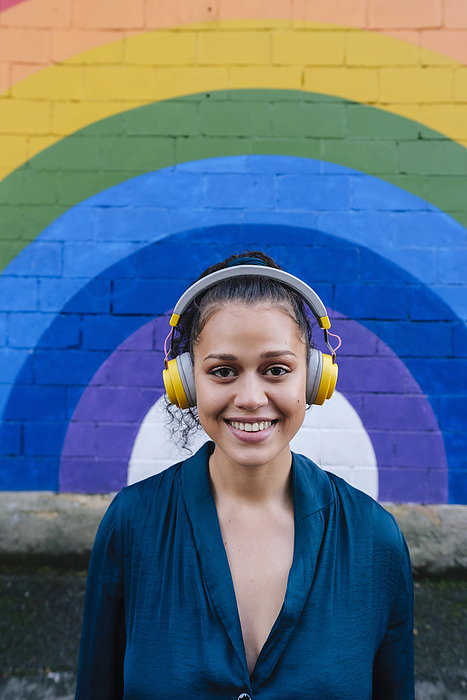 Happy young woman wearing wireless headphones in front of rainbow wall