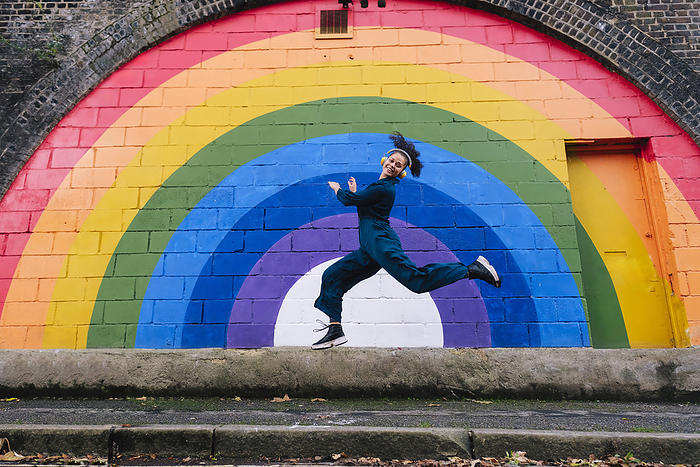 Happy young woman wearing wireless headphones and dancing in front of rainbow wall