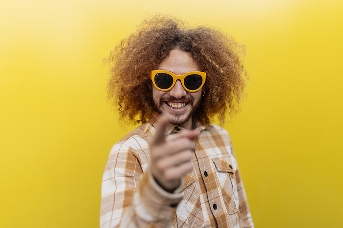 Happy man wearing sunglasses and pointing in front of yellow wall
