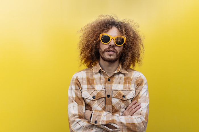 Man wearing sunglasses and standing with arms crossed in front of yellow wall