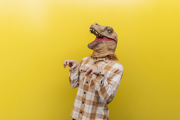 Man wearing dinosaur mask and gesturing in front of yellow wall